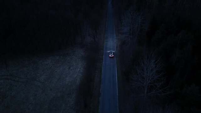 Car driving along straight road in dense forest at night filmed from drone with tracking movement and visible headlights