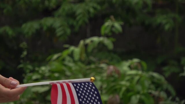 A hand waving a small US flag back and forth against a blurred background of green forest leaves.