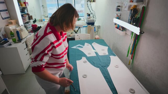 Woman carefully traces pattern onto fabric. She focuses intently, moving slowly with precision. Workshop space bright and organized with tools nearby. Footage ideal for fashion design, tailoring