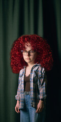 Girl with oversized red curly hair and glasses stands against dark green curtain. Concept of bold...