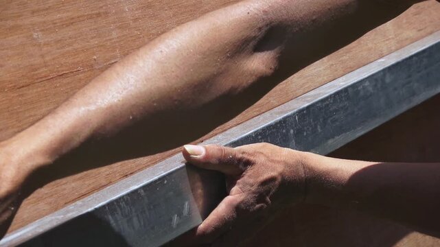 A worker's hands using a long aluminum straight edge and a pen to mark a cutting line on a large sheet of plywood.