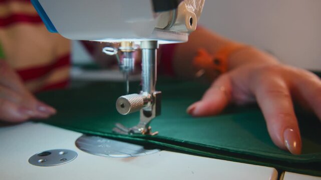 Hands guide green fabric under sewing machine needle. Close-up shows steady stitching motion. Fabric moves smoothly with precise control. Ideal for fashion, crafting, or textile industry projects