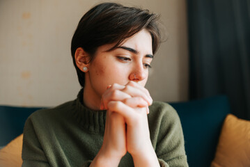 A young woman with short hair is sitting indoors with her hands clasped together, appearing thoughtful. The warm colors of the room create a cozy atmosphere around her reflective mood © Sergio