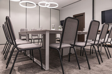 Fototapeta premium A spacious modern conference room features a long white table surrounded by grey chairs, clear glasses, small floral arrangements in vases, large windows with curtains, and stylish circular ceiling li