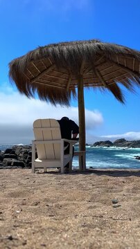 Vertical video of a young man sitting in a wooden chair under a thatched-roof palm tree at Punta Pite Beach on Chile's central coast