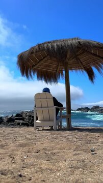 A vertical establishing view of a young man wearing a cap, sitting on a wooden chair under a parasol made of natural materials on the coast at Punta Pite Beach, Chile