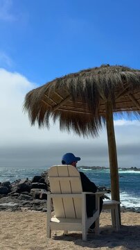 A vertical establishing view of a young man sitting on the shore at Punta Pite Beach on Chile&rsquo;s central coast