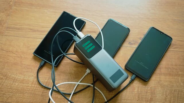Portable power bank charging three smartphones via multiple cables simultaneous charging setup. External battery powering several mobile devices at once. Multi phone charging process continuing with