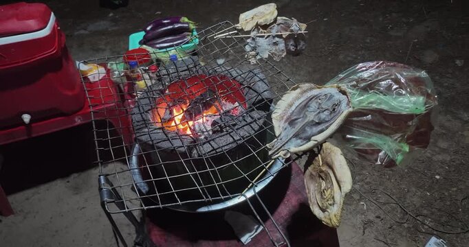close up of a traditional clay charcoal stove grilling dried fish at night with glowing embers and flying sparks.