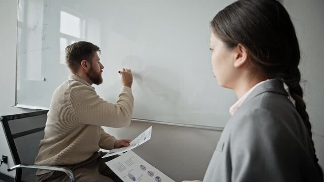 Asian woman with document looking at Caucasian man drawing schemes on wall whiteboard and listening to him while they preparing for meeting in office