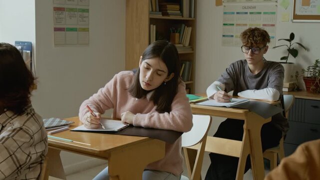 Full shot of motivated Caucasian male and female high school students sitting at desks at lesson, writing test, looking up at blackboard, to achieve good grades for trimester during academic year