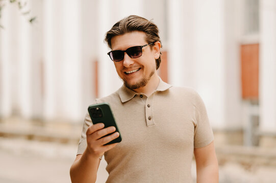Smiling Caucasian Man Checking Smartphone On City Sidewalk, Wearing Sunglasses And Beige Polo, Casual