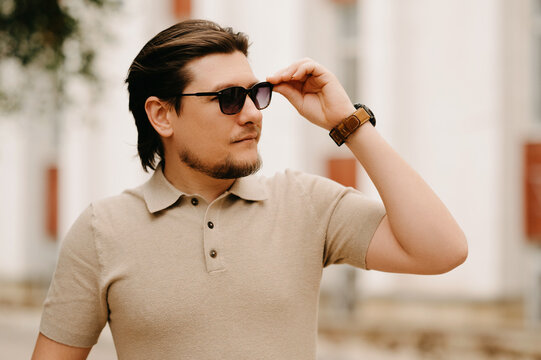 Pensive Man Lifting Sunglasses Beside Architectural Facade, Polo Shirt, Tiedback Hair, Wristwatch Content