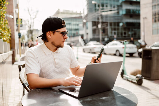 Young Man Using Laptop And Smartphone At Outdoor Cafe Table On Busy City Sidewalk, Wearing Cap And Sunglasses,