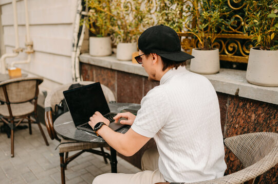 Focused Man Typing On Laptop At Quiet Courtyard Table Surrounded By Plants And Stone Wall, Wearing Cap