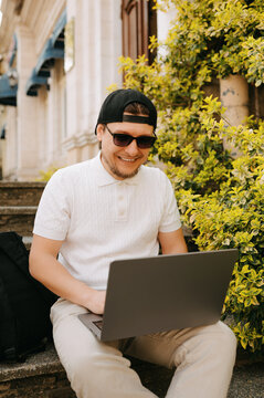 Focused Caucasian Man Typing On Laptop On Outdoor Bench Surrounded By Foliage, Wearing Backward Cap