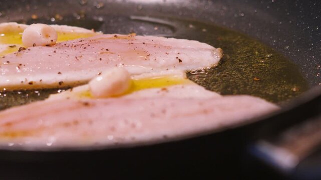 Two sea bass fillets in a hot pan cooking skin side down in bubbling olive oil. White fish cooked with garlic. Healthy omega-3 rich, high protein food. 4K.