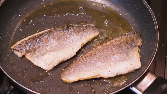 Top down view of two sea bass fillets in a hot frying pan in bubbling olive oil. White fish with crispy golden skin. Healthy omega-3 rich, high protein food. 4K.