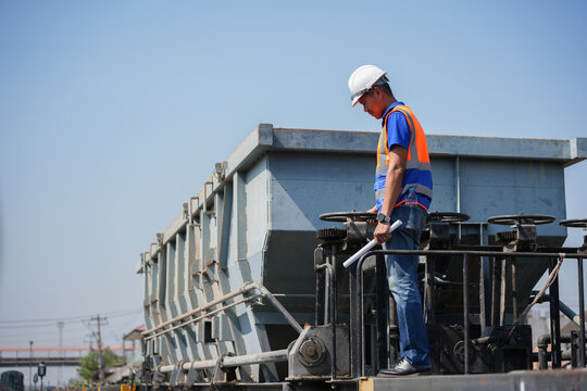 Professional railway engineer standing on freight car and inspecting cargo operations, Technician in safety vest and hardhat checking industrial hopper wagon at rail yard