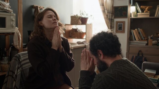Low angle shot of poor married couple praying while facing each other, supoorting one another through difficult times in small apartment