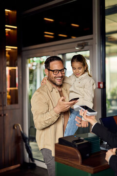 Smiling Father With Daughter Checking In At Hotel Reception Using Mobile Payment