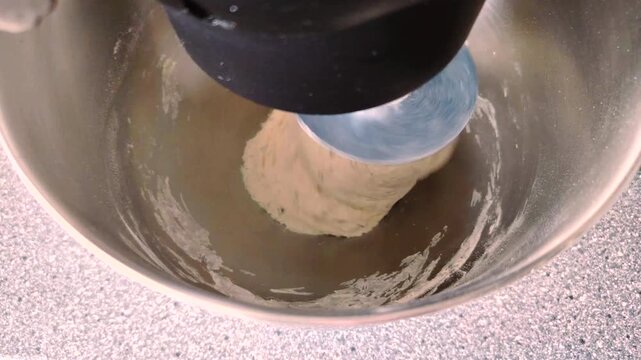 Planetary mixer kneading bread dough with a spiral hook in a metal bowl.