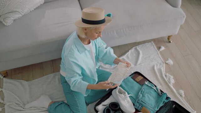Stylish elderly woman packing suitcase for summer trip, putting clothes and map. Happy female traveler dreaming about tourism and traveling in unknown city, traditional paper map for navigation
