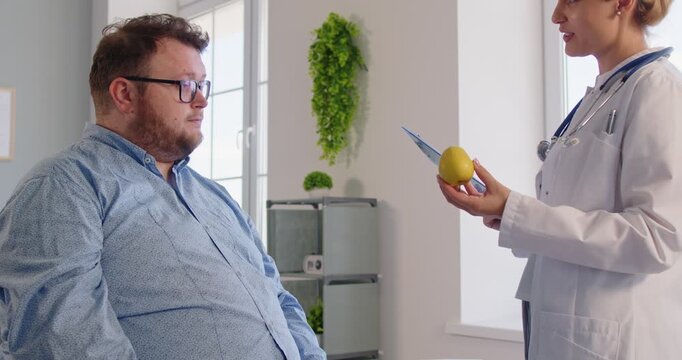 Doctor provides nutrition counseling, handing an apple to fat overweight man patient with obesity during a clinic visit to encourage diet change. Nutritionist gives apple as healthy symbol.