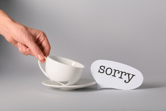 Female hand with broken tea cup and word SORRY on grey background