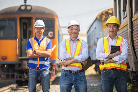 Confident team of Asian railway engineers and technicians standing at train station, Group of happy industrial workers and inspectors supervising train operations in freight depot
