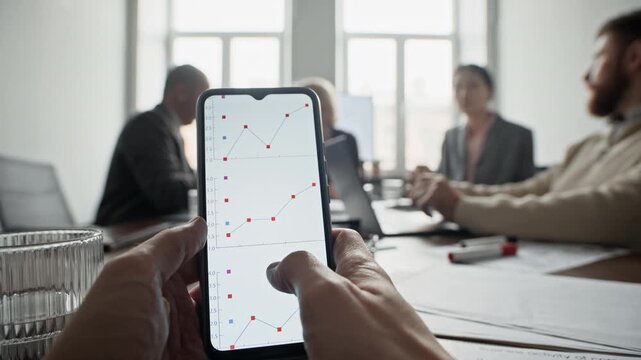 POV of unrecognizable man looking at smartphone screen and examining graphics while his colleagues communicating in blurred background