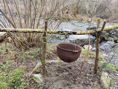 Old empty tourist cauldron suspended above campfire area on riverbank. Metal pot suspended from makeshift wooden crossbar mounted on slingshots. Close-up