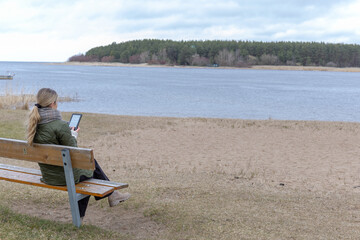 Fototapeta premium Woman sitting on bench by lake using tablet, remote work and reading outdoors, calm lifestyle and digital lifestyle concept