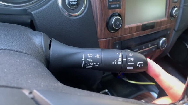 Finger switching windshield wiper control lever in car close-up, adjusting cleaning mode and activating system for rain visibility, demonstrating driving safety control, vehicle interior functionality
