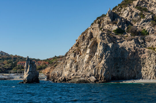 Narrow rock  rising above sea, Sail Rock in Simeiz sits against sheer cliff with coastal building and greenery in the background under clear blue sky. Crimea Yalta, Crimea &ndash; October 11, 2014
