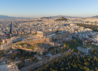 Fototapeta premium High-angle aerial image of the Parthenon atop the Acropolis hill, overlooking the vast urban sprawl of Athens and Lycabettus Hill at sunset.