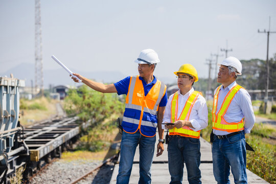 Team of Asian engineers pointing and discussing logistics plans at railway freight station, Group of railway technicians and managers in safety vests planning maintenance at rail yard