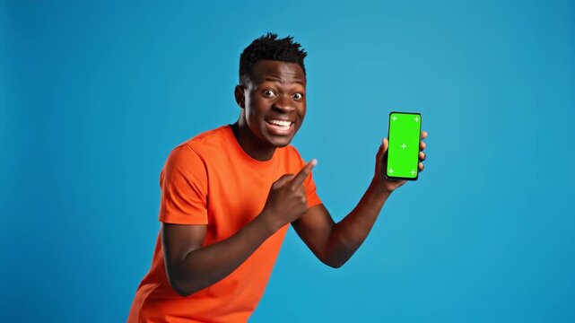 Happy young African American man in orange t-shirt shows phone with chroma key screen for mock-up. He points and expresses excitement, perfect for advertising.