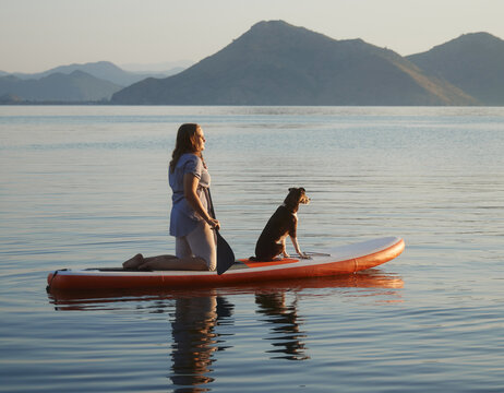 A woman stands with dog on an orange board floating quietly. Still lake water surrounds them beneath mountain views.