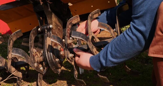 Close-up view of a farmer's hands securing the rotary tiller blades on a two-wheel tractor with a metal pin before starting fieldwork, highlighting agricultural machinery maintenance.