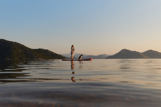 A woman stands with dog on a paddle board during dusk. The scene is quiet with smooth water and distant mountains.