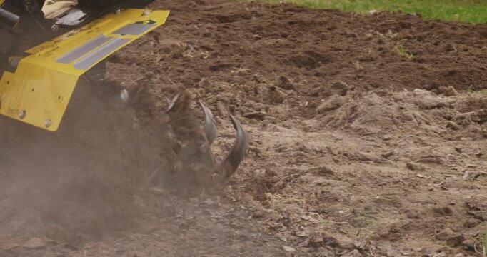 Farmer using a yellow motorized tiller to plow the land in a garden, loosening the soil before planting crops. Agricultural machinery preparing the field for the upcoming spring season.