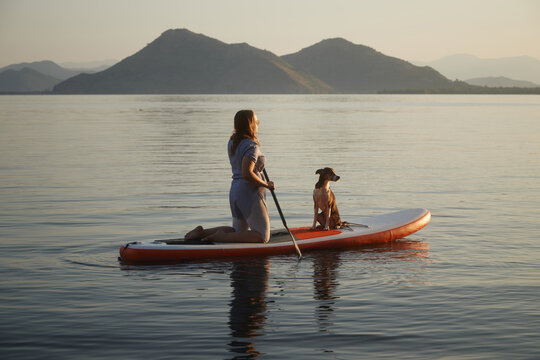 A woman stands tall on a board with a dog. The group floats together over smooth reflective water.