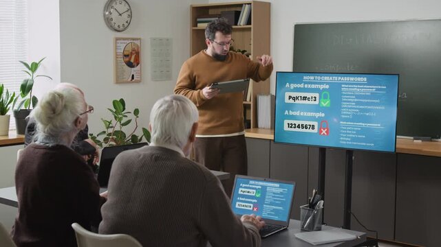 Male teacher giving lecture on digital safety in computer class for seniors and pointing at screen with password protection examples explaining internet scams and hackers, copy space