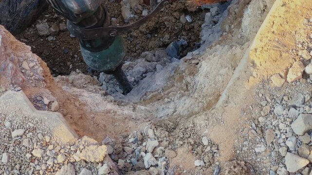 Close-up view of drilling machine bit penetrating soil and rock inside dug pit, rough textures and loose earth visible in a construction or excavation outdoor setting