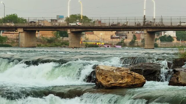 Close view of river rapids cascading over rocks with strong water flow, textured rock formation in foreground and bridge in background under soft natural daylight