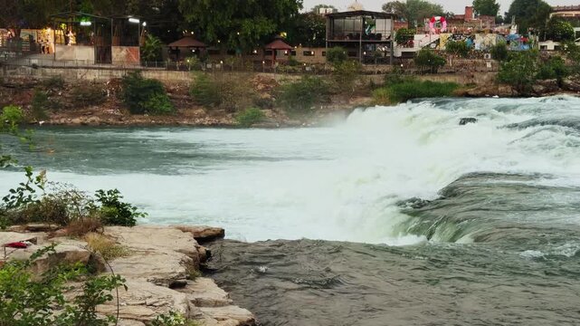 View of water cascading over rocky edge forming a waterfall at Eco Park Rewa, mist rising with riverside structures and trees in background under soft evening light