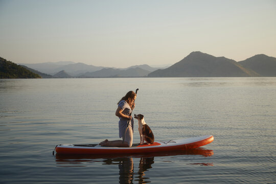 A woman stands with a dog while paddling across a flat lake. The reflections create a symmetrical and quiet view.