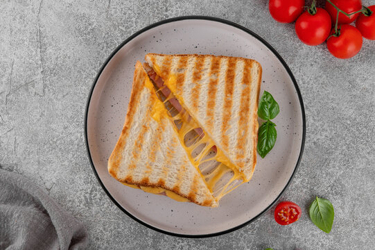 Grilled ham and cheese sandwich, cut in half with melted cheese stretching between the slices on a plate on a gray concrete background. Top view.