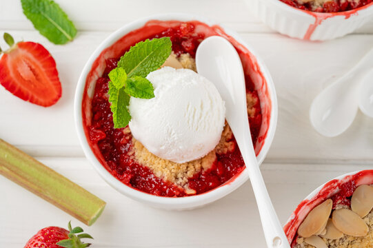 Rhubarb and strawberry crumble with a scoop of ice cream on top in a baking form on a white wooden background. Top view